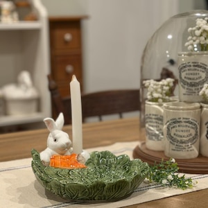 Może przedstawiać: Ceramiczny świecznik wielkanocny z białym królikiem trzymającym marchewkę, umieszczony w zielonej misce w kształcie sałaty. Biała świeca znajduje się pośrodku. Świecznik stoi na stole z kremowym bieżnikiem.
