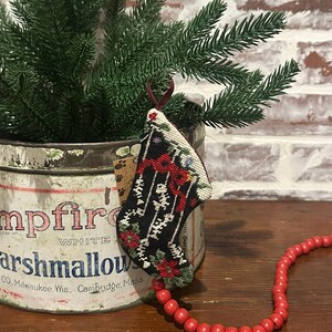 Puede incluir: Un calcetín navideño en miniatura con un fondo negro y bordados florales rojos y blancos. El calcetín cuelga de un pequeño árbol de hoja perenne en una lata de malvaviscos vintage. Un collar de cuentas de madera rojas está cerca.