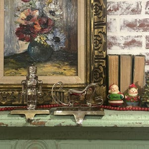 Puede incluir: Una pintura al óleo de flores en un jarrón se exhibe sobre una repisa decorada para Navidad. Un cascanueces plateado y unos sujetadores de medias en forma de trineo se encuentran en la repisa, junto con pequeñas figuras navideñas y cuentas rojas.