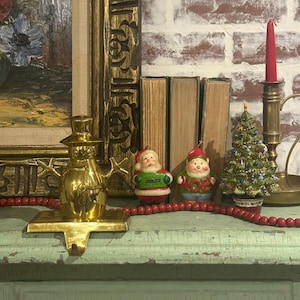 Peut inclure: Un support de bas de Noël en forme de bonhomme de neige doré, une guirlande de perles rouges, deux petits ornements de Père Noël et un sapin de Noël miniature. Une bougie rouge dans un bougeoir en laiton. Décoration de Noël.