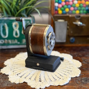 May include: A vintage pencil sharpener with a dark wood grain barrel and silver accents. The sharpener sits on a black base and has a silver crank handle. It rests on a cream-colored, crocheted doily. The background includes a green license plate and a gumball machine.