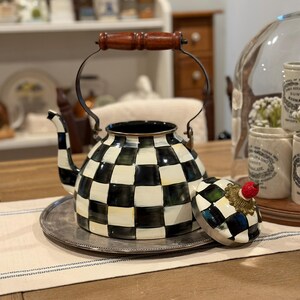 May include: A decorative black and white checkered teapot with a wooden handle and a silver-colored metal tray. The lid, adorned with a red fruit-shaped knob, sits beside the teapot. The teapot is a kitchenware item.
