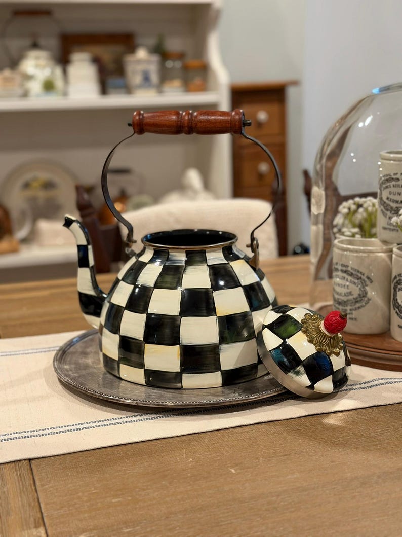 May include: A decorative black and white checkered teapot with a wooden handle and a silver-colored metal tray. The lid, adorned with a red fruit-shaped knob, sits beside the teapot. The teapot is a kitchenware item.