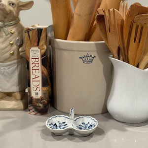 Può includere: Un piccolo piatto in ceramica per sale e pepe con un motivo floreale blu. Un grande vaso beige con mattarelli e utensili di legno sullo sfondo. È visibile anche un libro di cucina intitolato "Breads".