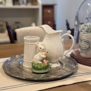 Puede incluir: Una jarra de cerámica blanca con superficie texturizada y asa, un pequeño vaso de cerámica blanca y una figura decorativa de conejito sosteniendo una zanahoria, todo dispuesto sobre una bandeja de metal plateado. La figura del conejito está sobre una base verde con flores rosas.