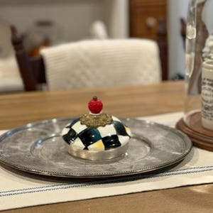 May include: A decorative ceramic jar lid with a black and white checkered pattern, topped with a red sphere and gold-colored accents. The lid sits on a silver-colored plate.