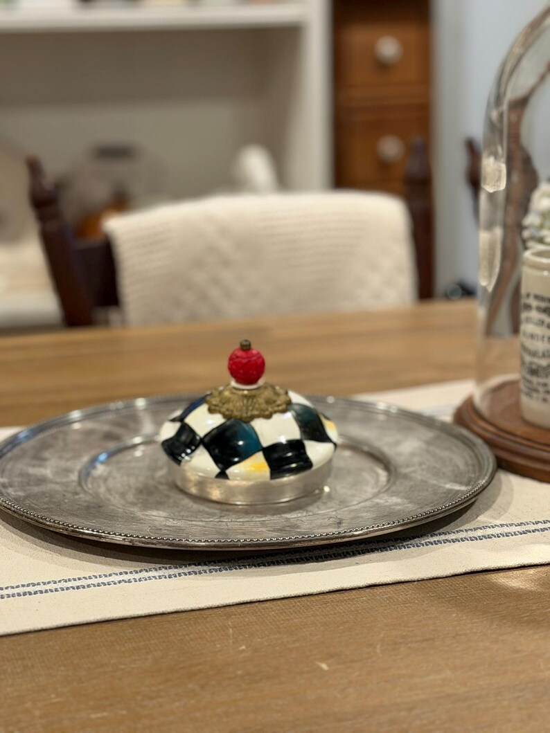 May include: A decorative ceramic jar lid with a black and white checkered pattern, topped with a red sphere and gold-colored accents. The lid sits on a silver-colored plate.