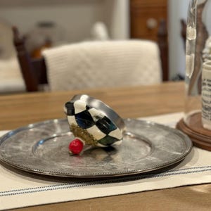 May include: A decorative, silver-colored plate with a beaded edge holds a black, white, and green checkered object with a gold base and a small red accent. The plate rests on a striped cloth.