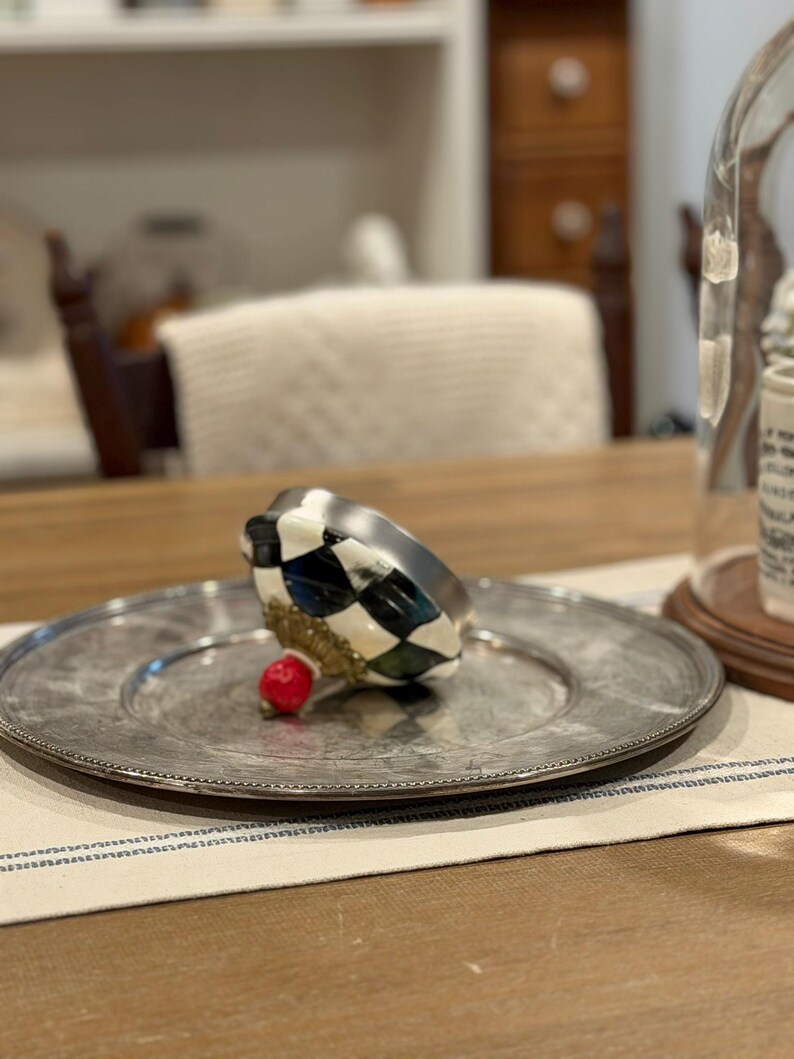 May include: A decorative, silver-colored plate with a beaded edge holds a black, white, and green checkered object with a gold base and a small red accent. The plate rests on a striped cloth.