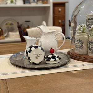 May include: A collection of decorative kitchenware on a silver tray. Includes a white pitcher with a dotted design, a small creamer, and a black and white checkered container with a red apple-shaped topper. A glass cloche with jars is in the background.
