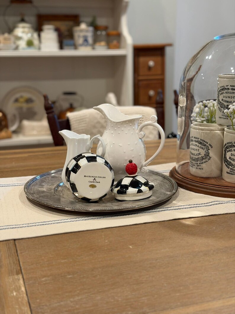 May include: A collection of decorative kitchenware on a silver tray. Includes a white pitcher with a dotted design, a small creamer, and a black and white checkered container with a red apple-shaped topper. A glass cloche with jars is in the background.