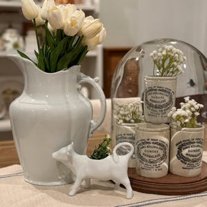 May include: A white ceramic pitcher filled with white tulips, a white cow-shaped planter with greenery, and several small vintage marmalade jars with white flowers. The jars are on a wooden tray, and the scene is set on a striped tablecloth.