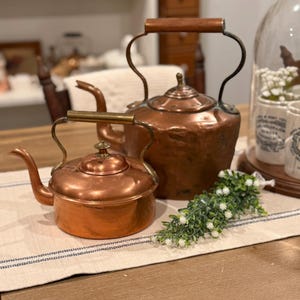 May include: Two antique copper teapots with brass handles and spouts sit on a wooden table. The larger teapot has a wooden handle. A sprig of white flowers and greenery adds a decorative touch to the scene.