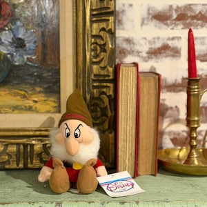 May include: A plush Grumpy dwarf doll from Disney's Snow White, wearing a red shirt, brown hat, and shoes. The doll has a white beard and a tag that reads "The Disney Store". The doll is sitting on a green surface.