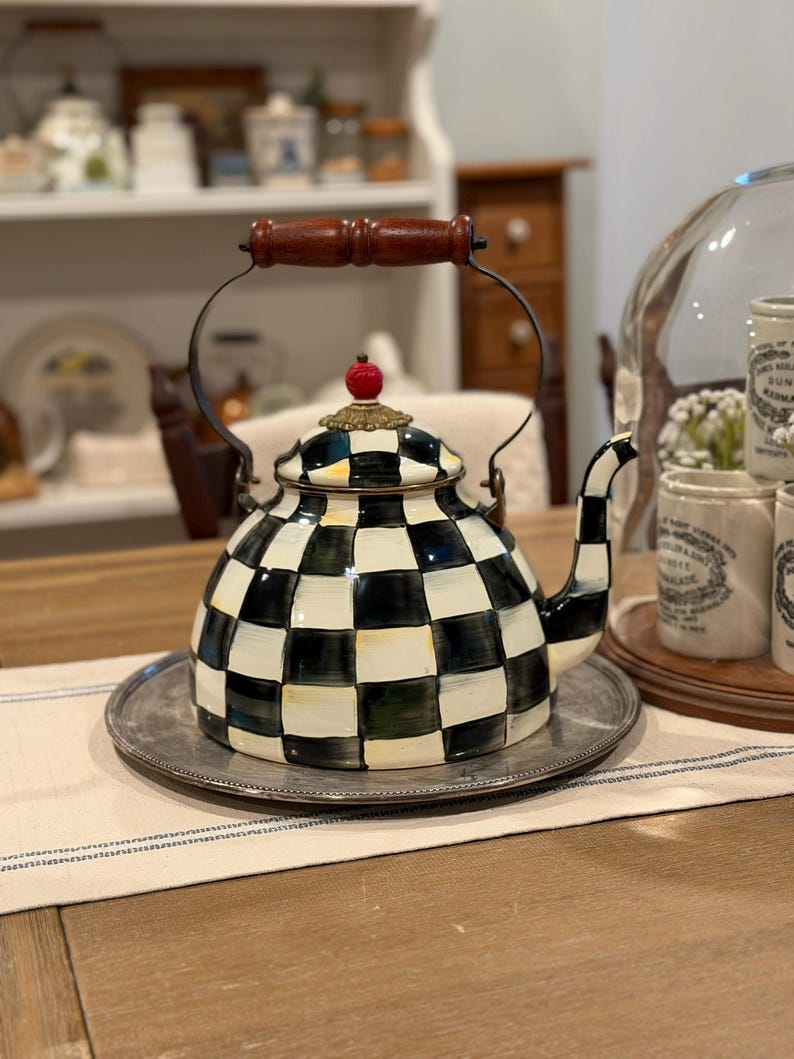 May include: A decorative black and white checkered teapot with a wooden handle and a red knob on top. The teapot sits on a silver tray, showcasing a classic design. The teapot is a kitchenware item.