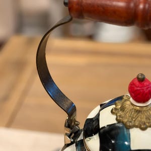 May include: Close-up of a decorative tea kettle with a black and white checkered pattern. The kettle features a wooden handle and a red and gold finial. The handle is made of dark wood and the metal parts are dark.