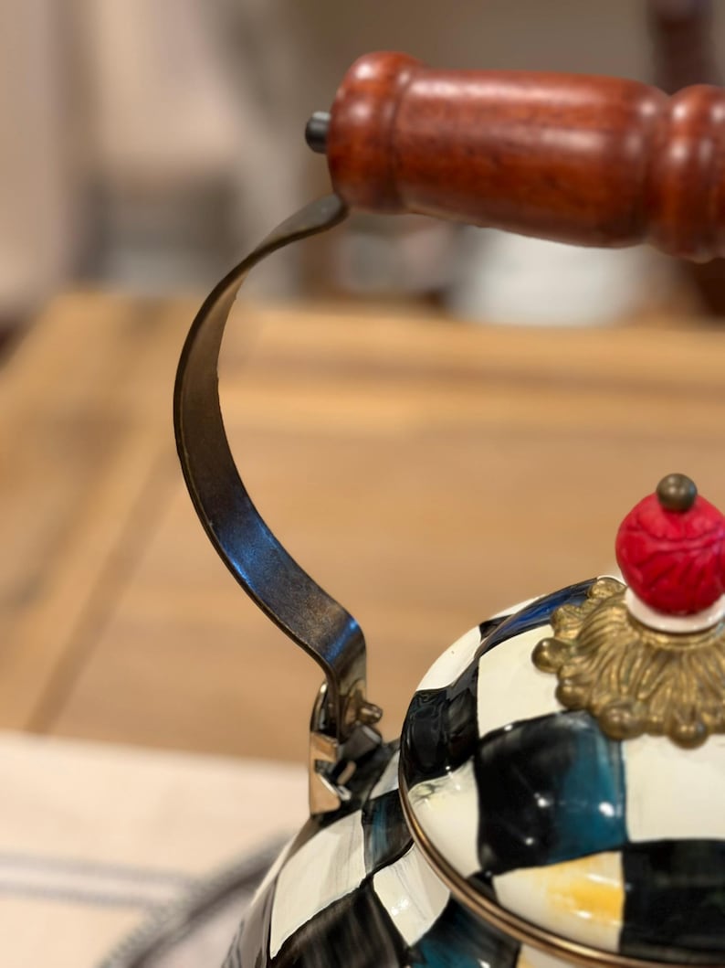 May include: Close-up of a decorative tea kettle with a black and white checkered pattern. The kettle features a wooden handle and a red and gold finial. The handle is made of dark wood and the metal parts are dark.
