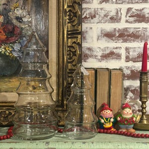 Peut inclure: Deux bocaux transparents en forme de sapin de Noël sont posés sur une surface verte vieillie. Une guirlande de perles rouges et deux figurines de gnomes sont devant un mur de briques. Une peinture encadrée et un bougeoir en laiton sont en arrière-plan.