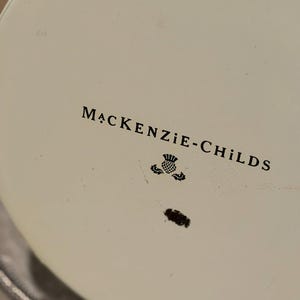 May include: Close-up of a white surface with the text "MACKENZIE-CHILDS" in black, along with a thistle emblem. The object has a black rim and a silver edge.