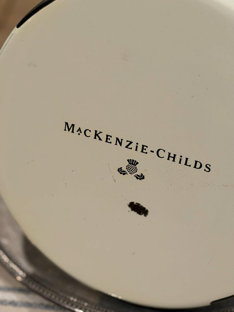 May include: Close-up of a white surface with the text "MACKENZIE-CHILDS" in black, along with a thistle emblem. The object has a black rim and a silver edge.