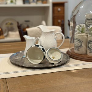 May include: A collection of white ceramic pitchers and a black and white checkered jar with a lid on a silver tray. A larger white pitcher with a dotted pattern is in the background. The scene is set on a wooden table.