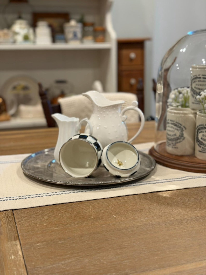 May include: A collection of white ceramic pitchers and a black and white checkered jar with a lid on a silver tray. A larger white pitcher with a dotted pattern is in the background. The scene is set on a wooden table.
