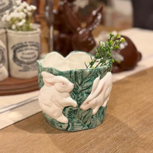 May include: A ceramic planter with a green base and white interior. The planter features white rabbit figures and green leaf designs. The planter is filled with greenery and is set on a wooden surface.