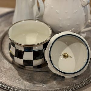May include: A decorative ceramic sugar bowl with a black and white checkered pattern, silver trim, and a matching lid. The lid is white with a blue trim. Two white ceramic pitchers are in the background, all on a silver tray.