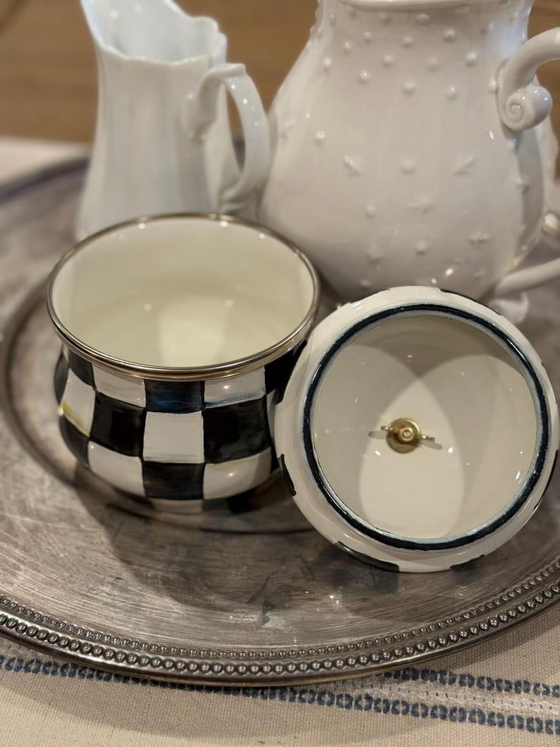 May include: A decorative ceramic sugar bowl with a black and white checkered pattern, silver trim, and a matching lid. The lid is white with a blue trim. Two white ceramic pitchers are in the background, all on a silver tray.