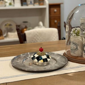 May include: A decorative ceramic jar lid with a black and white checkered pattern, topped with a gold-colored accent and a red sphere. The lid sits on a silver-colored plate, placed on a table runner.