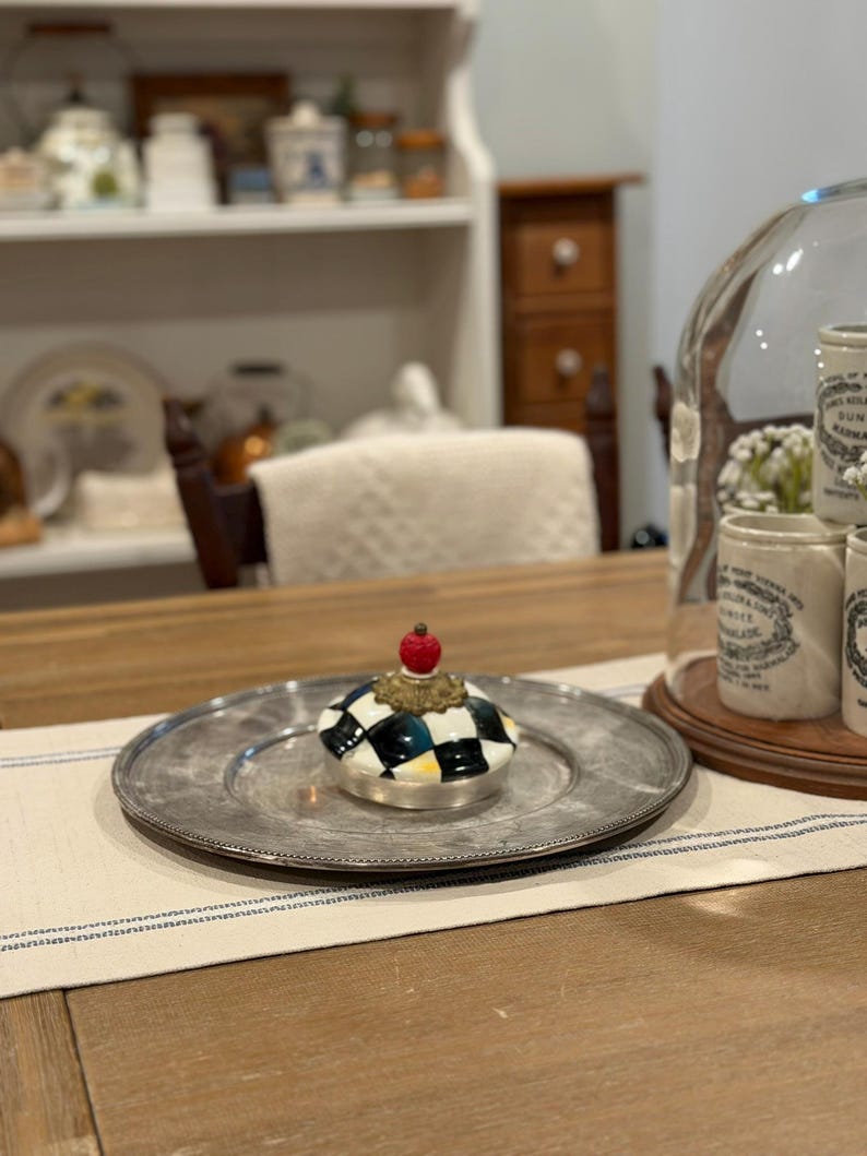 May include: A decorative ceramic jar lid with a black and white checkered pattern, topped with a gold-colored accent and a red sphere. The lid sits on a silver-colored plate, placed on a table runner.