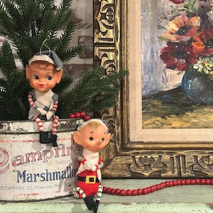 Peut inclure: Deux ornements de Noël vintage en forme d'elfes. L'un est assis sur une boîte de conserve avec l'inscription "Campfire Marshmallows". L'autre porte un costume rouge et blanc avec une ceinture noire. Une guirlande de perles rouges et une peinture encadrée sont en arrière-plan.