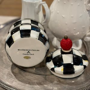May include: A black and white checkered ceramic jar with a matching lid topped with a red cherry. The jar is labeled "MacKenzie-Childs Courtly Check". Two white pitchers are in the background, all on a silver tray.