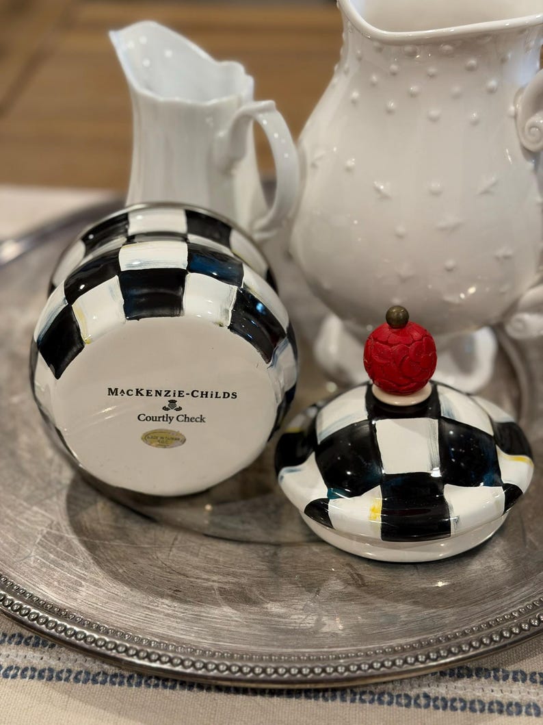 May include: A black and white checkered ceramic jar with a matching lid topped with a red cherry. The jar is labeled "MacKenzie-Childs Courtly Check". Two white pitchers are in the background, all on a silver tray.