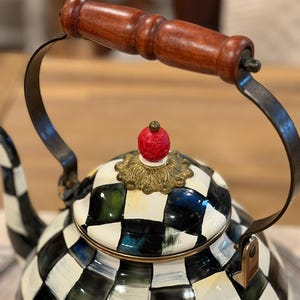 May include: A decorative tea kettle with a black and white checkered pattern. The handle is made of dark wood, and the lid features a red, ornate finial. The kettle's handle is made of dark metal.