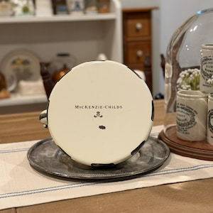 May include: A white teacup with black accents and the text "McKenzie-Childs" on the bottom, resting on a silver saucer. The cup is part of a set, with a matching design. The teacup is on a table with a blue and white striped runner.