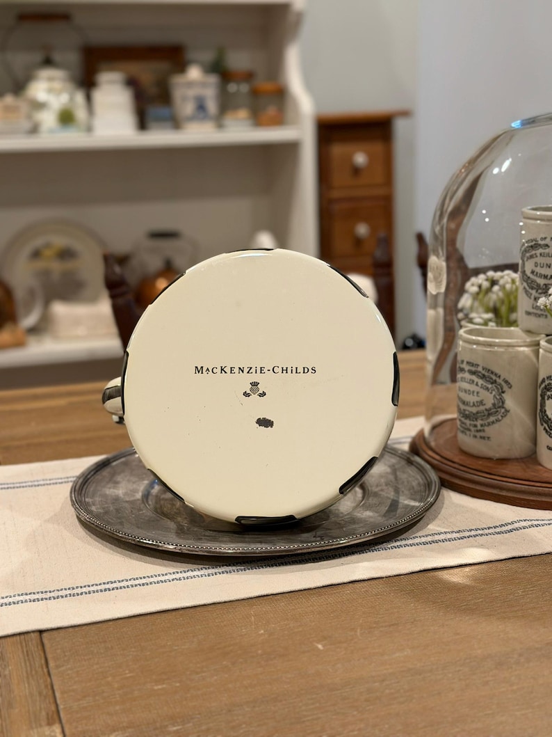 May include: A white teacup with black accents and the text "McKenzie-Childs" on the bottom, resting on a silver saucer. The cup is part of a set, with a matching design. The teacup is on a table with a blue and white striped runner.
