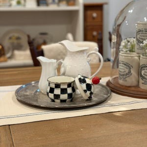 May include: A silver tray holds a small white pitcher, a larger white pitcher with a dotted texture, and a black and white checkered jar with a lid. A red cherry sits on the lid. The tray sits on a cream-colored runner.