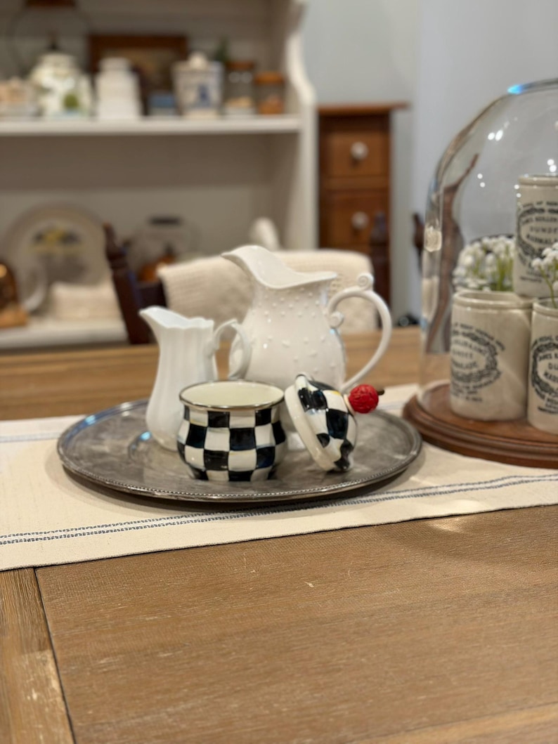 May include: A silver tray holds a small white pitcher, a larger white pitcher with a dotted texture, and a black and white checkered jar with a lid. A red cherry sits on the lid. The tray sits on a cream-colored runner.