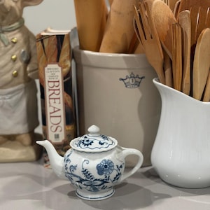 Könnte beinhalten: Eine kleine, weiße Porzellan-Teekanne mit blauen Blumenmustern und passendem Deckel. Die Teekanne steht vor einer Sammlung von Küchenutensilien, darunter Nudelrollen und Holzlöffel, sowie einem Keramiktopf.