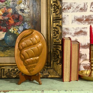 Puede incluir: Un molde decorativo ovalado de color cobre con un diseño en relieve, exhibido sobre un pequeño soporte de madera. El molde se encuentra frente a un cuadro enmarcado, libros y una pared de ladrillos.