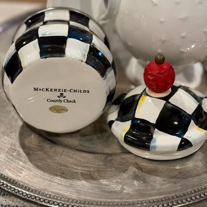 May include: A ceramic jar with a black and white checkered pattern, with the words "MacKenzie-Childs Courtly Check" printed on the bottom. The lid is off, revealing the inside of the jar. The lid has a red, textured knob.