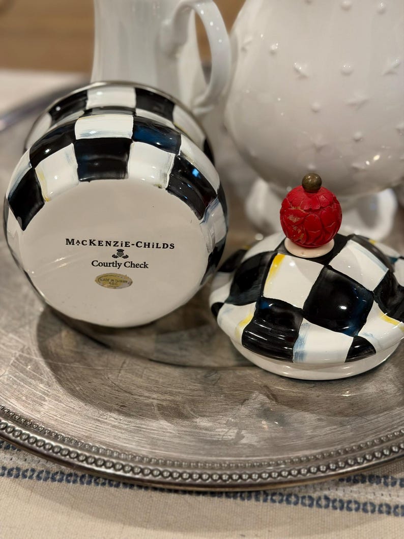 May include: A ceramic jar with a black and white checkered pattern, with the words "MacKenzie-Childs Courtly Check" printed on the bottom. The lid is off, revealing the inside of the jar. The lid has a red, textured knob.
