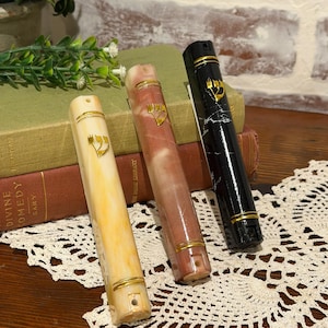 May include: Three decorative mezuzah cases in a row. One is cream-colored, one is pink and brown, and the third is black with white marbling. Each has a gold-colored emblem and gold bands. They are displayed on a table with books and lace.