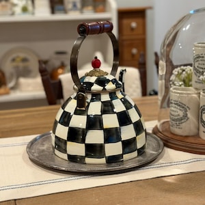 May include: A decorative black and white checkered teapot with a silver metal base. The teapot has a brown wooden handle and a red decorative top. The teapot is sitting on a silver tray on a table.