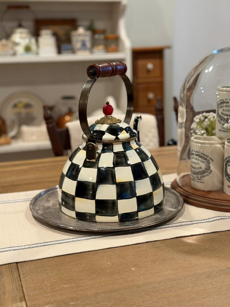 May include: A decorative black and white checkered teapot with a silver metal base. The teapot has a brown wooden handle and a red decorative top. The teapot is sitting on a silver tray on a table.