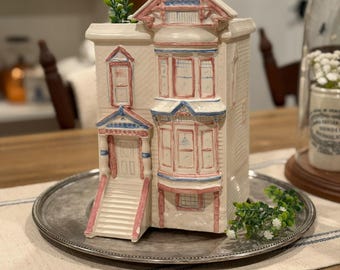 May include: A ceramic house-shaped planter with a detailed facade, painted in cream, pink, and blue. The planter sits on a silver tray and features a small green plant. The house has a staircase, windows, and a decorative roof.