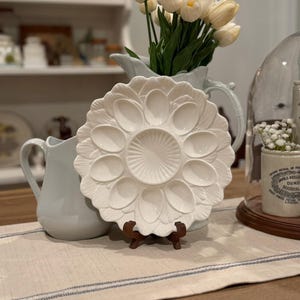 May include: A white ceramic oyster serving platter with 12 oval compartments and a central circular area, displayed on a wooden stand. The platter is surrounded by a light blue pitcher with white tulips and other decorative items.