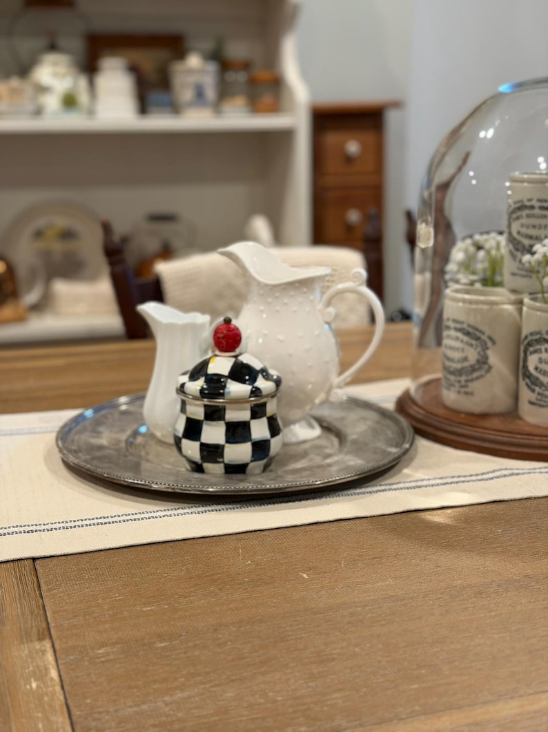 May include: A decorative arrangement featuring a silver tray holding a small white pitcher, a white dotted pitcher, and a black and white checkered jar with a red cherry-like top. The items are on a wooden table.