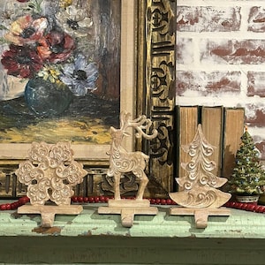 May include: Three beige Christmas stocking holders: a snowflake, a reindeer, and a Christmas tree. The holders are on a distressed green surface, with a red beaded garland. A framed painting and books are in the background.